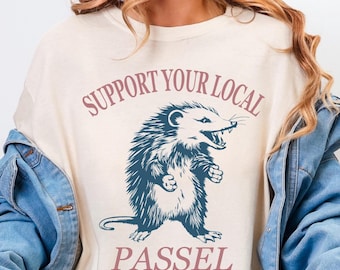 May include: Ivory-colored t-shirt featuring a detailed illustration of an opossum with the text "SUPPORT YOUR LOCAL PASSEL" in a pinkish-purple hue. The opossum is depicted with its mouth open, and the shirt is a relaxed fit.