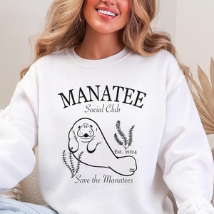 May include: White sweatshirt featuring a black graphic of a manatee with the text "MANATEE Social Club" and "Save the Manatees". The design includes the year "Est. 2024". The sweatshirt is a casual style.