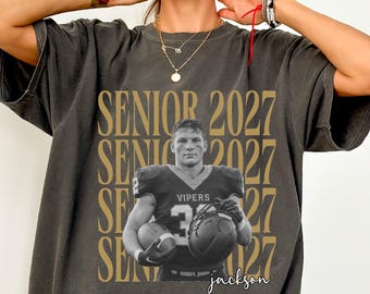 May include: A charcoal gray short-sleeve t-shirt featuring a graphic print of a football player in a uniform labeled Vipers. The text Senior 2027 is repeated in gold behind the player, and the name Jackson is written in script below the image.