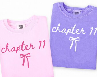 May include: Two crewneck sweatshirts, one blossom pink and one violet, are displayed. Each features the text "chapter 11" in a cursive font, along with a bow graphic. The sweatshirts are from Comfort Colors.