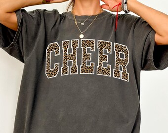 May include: A dark gray, oversized t-shirt with the word "CHEER" in a leopard print design. The text is outlined in white. The shirt is a relaxed fit and has short sleeves.