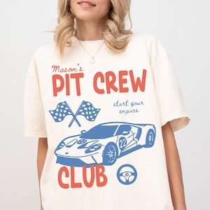 May include: Ivory-colored t-shirt with a graphic design featuring the text "Mason's PIT CREW CLUB" in red, a blue race car, crossed checkered flags, and a steering wheel. The phrase "start your engines" is also included.