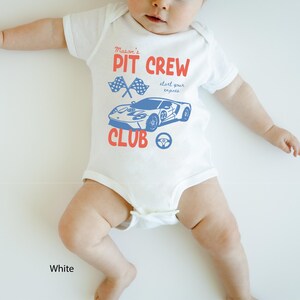 May include: White baby onesie with a red and blue graphic. The onesie features the text "Mason's PIT CREW CLUB" with a race car and checkered flags. The text "start your engines" is also included.