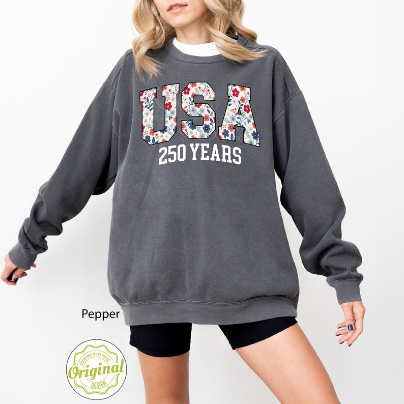 Puede incluir: Una sudadera gris oscuro con la palabra "USA" en un dise&ntilde;o floral y el texto "250 YEARS" en blanco. La sudadera es de cuello redondo y tiene mangas largas. La palabra "Pepper" est&aacute; impresa en la parte inferior izquierda.