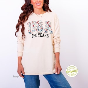 Puede incluir: Sudadera de manga larga color crema con el texto "USA 250 YEARS" en una fuente universitaria. Las letras "USA" est&aacute;n rellenas con un estampado floral en rojo, azul y blanco. La sudadera es de corte holgado.