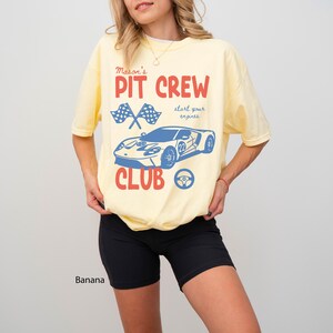 May include: A light yellow t-shirt with a graphic design featuring a blue race car, checkered flags, and the words "PIT CREW CLUB" in red. The shirt also includes the text "start your engines" in a script font. The word "Banana" is printed at the bottom.