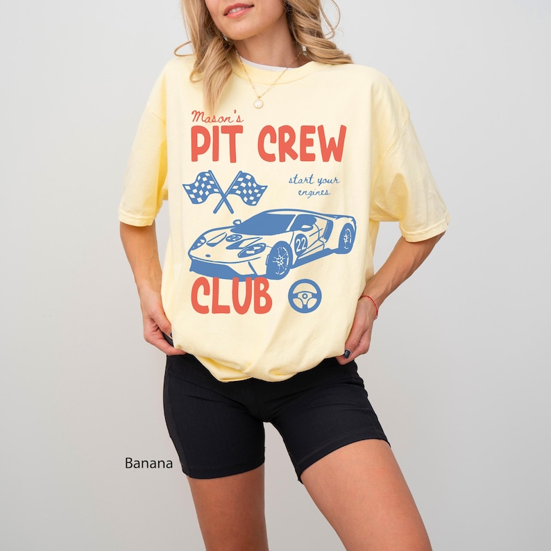May include: A light yellow t-shirt with a graphic design featuring a blue race car, checkered flags, and the words "PIT CREW CLUB" in red. The shirt also includes the text "start your engines" in a script font. The word "Banana" is printed at the bottom.