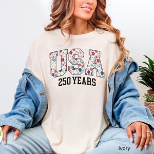 Puede incluir: Camiseta de color marfil con la palabra "USA" en un estampado floral y el texto "250 YEARS" en negro. La camiseta se lleva con una chaqueta vaquera azul y vaqueros azul claro. La imagen tambi&eacute;n incluye plantas en macetas.