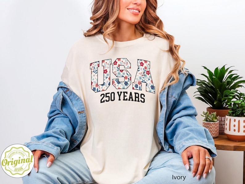 Puede incluir: Camiseta de color marfil con la palabra "USA" en un estampado floral y el texto "250 YEARS" en negro. La camiseta se lleva con una chaqueta vaquera azul y vaqueros azul claro. La imagen tambi&eacute;n incluye plantas en macetas.