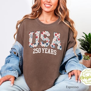 Puede incluir: Camiseta color espresso con el texto "USA 250 YEARS" en un dise&ntilde;o floral. La camiseta es de corte holgado y se lleva con una chaqueta vaquera. El dise&ntilde;o tiene un tema patri&oacute;tico.