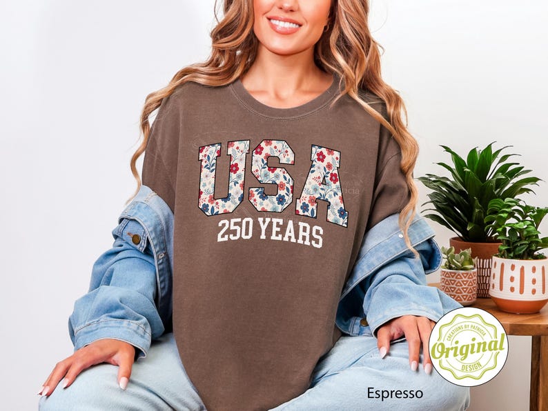 Puede incluir: Camiseta color espresso con el texto "USA 250 YEARS" en un dise&ntilde;o floral. La camiseta es de corte holgado y se lleva con una chaqueta vaquera. El dise&ntilde;o tiene un tema patri&oacute;tico.