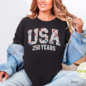Puede incluir: Sudadera negra de cuello redondo con la palabra "USA" en estampado floral y el texto "250 YEARS" en blanco. La sudadera forma parte de una colecci&oacute;n de ropa.