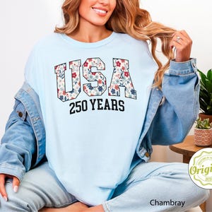 Puede incluir: Sudadera azul claro con la palabra "USA" en un estampado floral y el texto "250 YEARS" en negro. La modelo lleva una chaqueta vaquera azul claro y vaqueros azules claros.