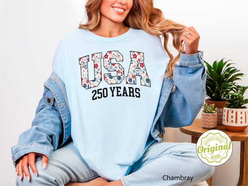 Puede incluir: Sudadera azul claro con la palabra "USA" en un estampado floral y el texto "250 YEARS" en negro. La modelo lleva una chaqueta vaquera azul claro y vaqueros azules claros.