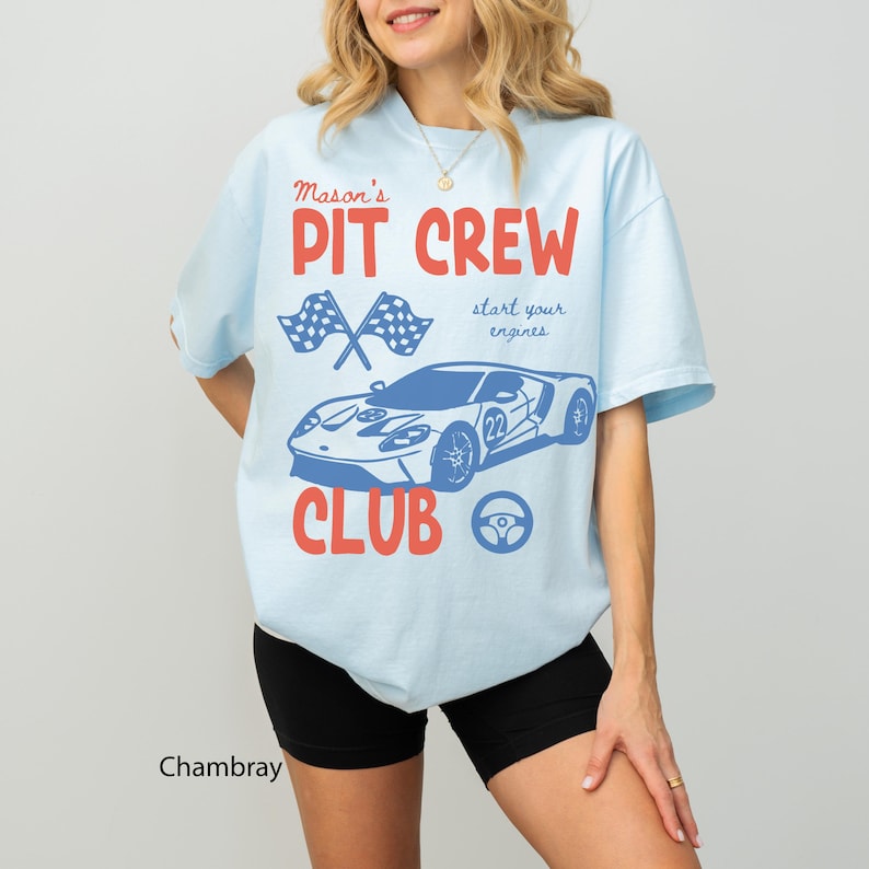 May include: Light blue t-shirt with the text "Mason's PIT CREW CLUB" in red and blue. The design includes crossed checkered flags, a race car, and a steering wheel. The shirt is made of a soft material.