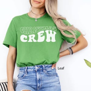 May include: A green t-shirt featuring the words "CHRISTMAS CREW" in a white, retro font. The shirt is a solid green colour and is worn with blue denim jeans. The word "Leaf" is printed on the bottom right of the image.