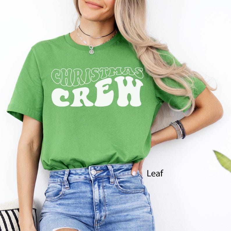 May include: A green t-shirt featuring the words "CHRISTMAS CREW" in a white, retro font. The shirt is a solid green colour and is worn with blue denim jeans. The word "Leaf" is printed on the bottom right of the image.