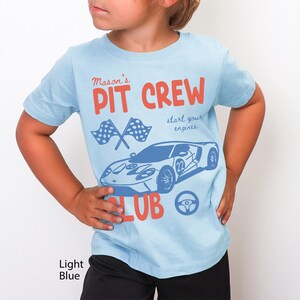 May include: Light blue t-shirt with a car graphic and the words "Mason's Pit Crew Club". The design includes checkered flags, a steering wheel, and the phrase "start your engines".