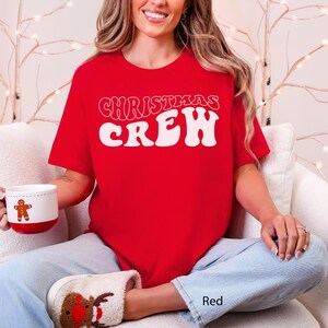May include: A red t-shirt with the words "CHRISTMAS CREW" in white, retro-style lettering. The person is wearing light blue jeans and reindeer slippers. A white mug with a gingerbread man is held in the hand.