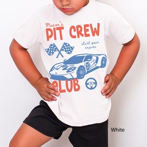 May include: White t-shirt with a blue and red graphic design. The design includes the text "Mason's PIT CREW CLUB" with a race car, checkered flags, and a steering wheel. The shirt is worn by a person wearing black shorts.