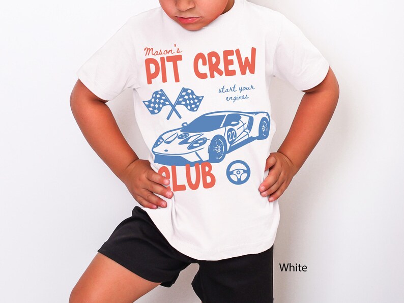 May include: White t-shirt with a blue and red graphic design. The design includes the text "Mason's PIT CREW CLUB" with a race car, checkered flags, and a steering wheel. The shirt is worn by a person wearing black shorts.