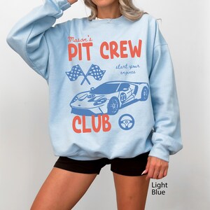 May include: Light blue sweatshirt with a graphic design featuring a race car, checkered flags, and the words "Mason's Pit Crew Club." The design also includes the phrase "start your engines." The sweatshirt is a light blue color.