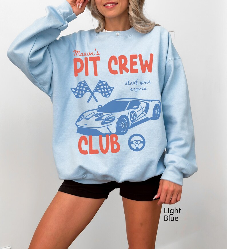 May include: Light blue sweatshirt with a graphic design featuring a race car, checkered flags, and the words "Mason's Pit Crew Club." The design also includes the phrase "start your engines." The sweatshirt is a light blue color.