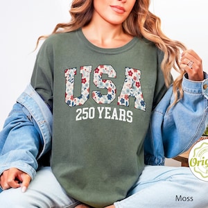 Puede incluir: Camiseta verde musgo con la palabra "USA" en un estampado floral y el texto "250 YEARS" en blanco. La camiseta forma parte de una colecci&oacute;n de ropa.