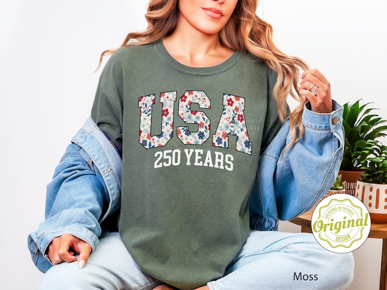 Puede incluir: Camiseta verde musgo con la palabra "USA" en un estampado floral y el texto "250 YEARS" en blanco. La camiseta forma parte de una colecci&oacute;n de ropa.
