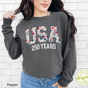Puede incluir: Sudadera de manga larga gris oscuro con la palabra "USA" en un estampado floral y el texto "250 YEARS" en blanco. La sudadera es de estilo casual, adecuada para el uso diario.