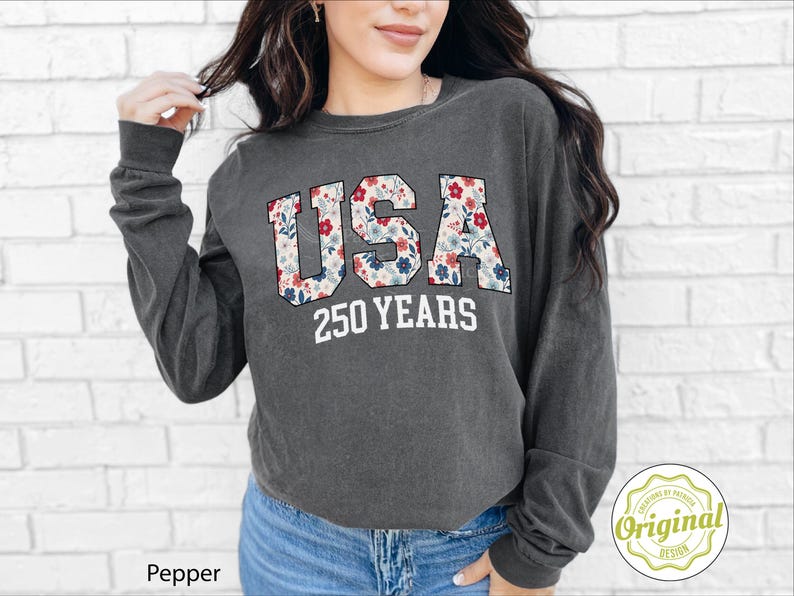Puede incluir: Sudadera de manga larga gris oscuro con la palabra "USA" en un estampado floral y el texto "250 YEARS" en blanco. La sudadera es de estilo casual, adecuada para el uso diario.