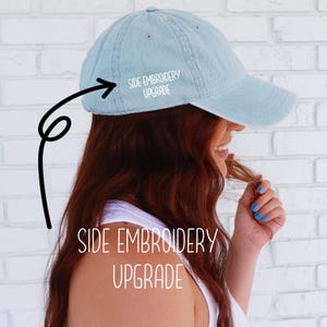Puede incluir: Gorra de béisbol azul claro con el texto "SIDE EMBROIDERY UPGRADE" en el lateral y en la parte inferior. La gorra tiene una visera curva y la lleva una persona con pelo rojo.