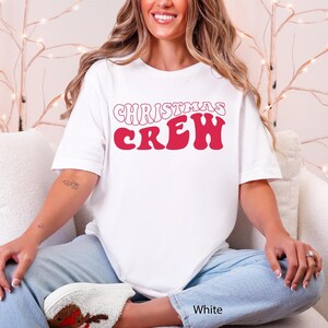 May include: White t-shirt featuring the words "CHRISTMAS CREW" in red, retro lettering. The casual, short-sleeved top is ideal for festive occasions. The model is wearing light blue jeans.