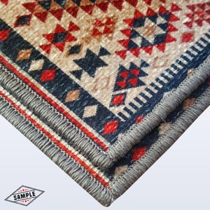 May include: Close-up of a corner of a patterned rug. The rug features a beige background with red, navy blue, and brown geometric designs. A gray border with fringe edges the rug, and a red stripe runs along the edge.