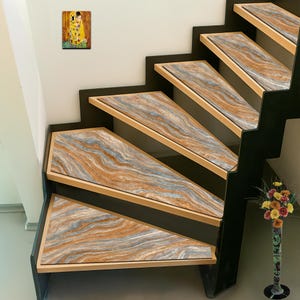 May include: A staircase with steps featuring a marble-like design in shades of brown, gray, and beige. The steps are framed with light wood and set against a black structure. A framed artwork hangs on the wall, and a vase of flowers is visible.