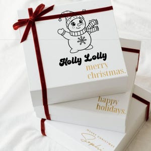 Op de afbeelding: Gestapelde witte geschenkdozen, versierd met rood fluwelen lint. De bovenste doos heeft een sneeuwpop illustratie en de tekst "Holly Lolly merry christmas." Andere dozen tonen "happy holidays" en "merry christmas Sophie."
