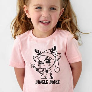 Op de afbeelding: Lichtroze T-shirt met een cartoon rendier met een kerstmuts en een toverstaf. Onder het rendier staat in zwarte letters "JINGLE JUICE".
