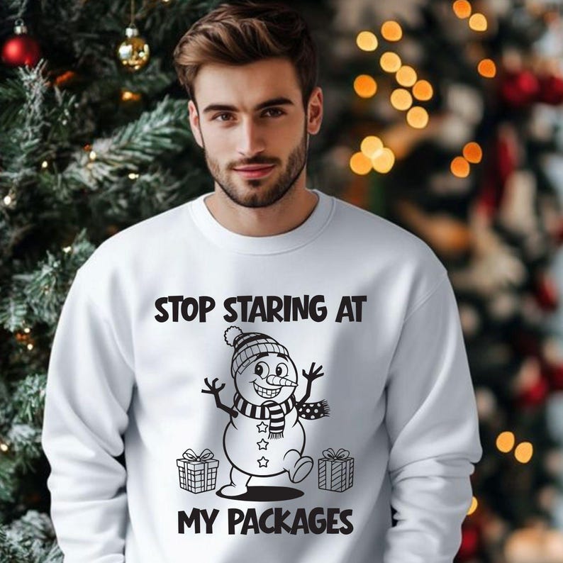 Op de afbeelding: Witte sweatshirt met de tekst "STOP STARING AT MY PACKAGES" en een cartoon sneeuwpop. De sneeuwpop draagt een hoed en sjaal, met cadeaudozen aan weerszijden. Op de achtergrond staat een kerstboom.
