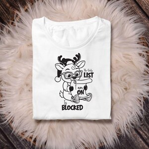 Op de afbeelding: Wit t-shirt met een afbeelding van een cartoon rendier met een bril en een kerstmuts, met een scroll. De tekst op de scroll luidt "The Only List I am On Is BLOCKED."