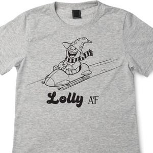 Op de afbeelding: Lichtgrijs t-shirt met een zwart-wit illustratie van een sneeuwpop met een heksenhoed en sjaal, die slee rijdt. De tekst "Lolly AF" staat onder de illustratie.