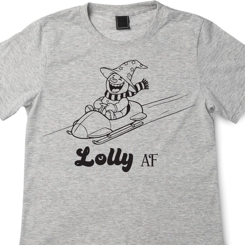 Op de afbeelding: Lichtgrijs t-shirt met een zwart-wit illustratie van een sneeuwpop met een heksenhoed en sjaal, die slee rijdt. De tekst "Lolly AF" staat onder de illustratie.