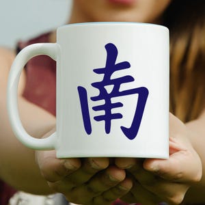 May include: White ceramic mug with a blue interior rim and a large blue Chinese character on the front. The mug has a curved handle and is held in both hands.