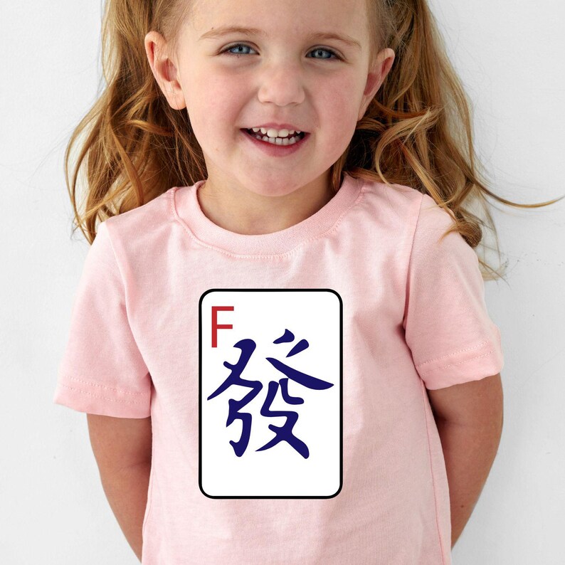 May include: A light pink t-shirt featuring a white rectangle with a black border. Inside the rectangle is a blue Chinese character and the letter "F" in red. The shirt is worn by a young child.