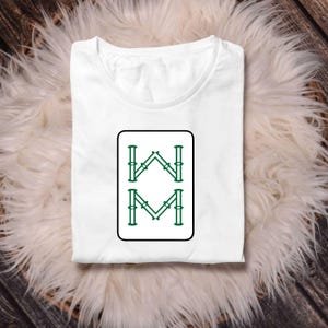 May include: White t-shirt with a crew neck, featuring a rectangular graphic with green and black accents. The graphic displays the letters "W" and "M" in a stylized design. The shirt is folded and rests on a fluffy, white surface.