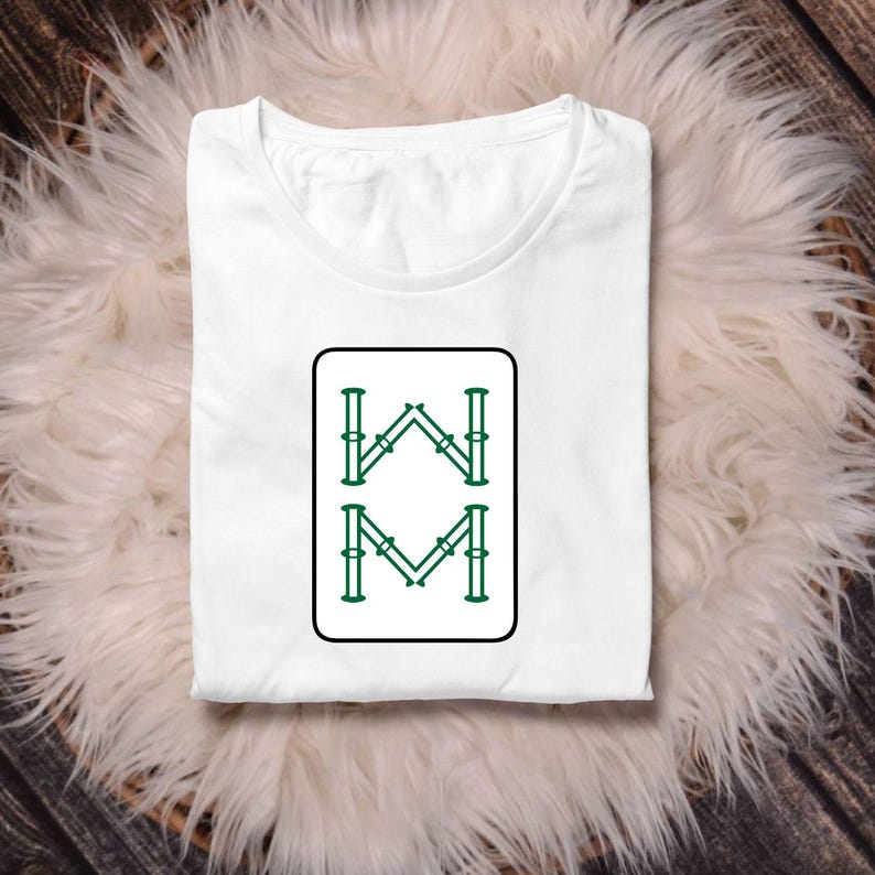 May include: White t-shirt with a crew neck, featuring a rectangular graphic with green and black accents. The graphic displays the letters "W" and "M" in a stylized design. The shirt is folded and rests on a fluffy, white surface.