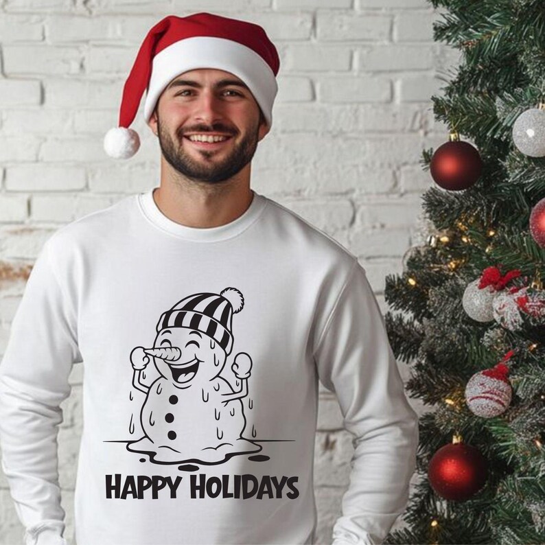 Op de afbeelding: Wit sweatshirt met een zwart-witte cartoon sneeuwpop afbeelding en de tekst "HAPPY HOLIDAYS". De persoon op de afbeelding draagt een rood-witte kerstmuts. Op de achtergrond staat een kerstboom.