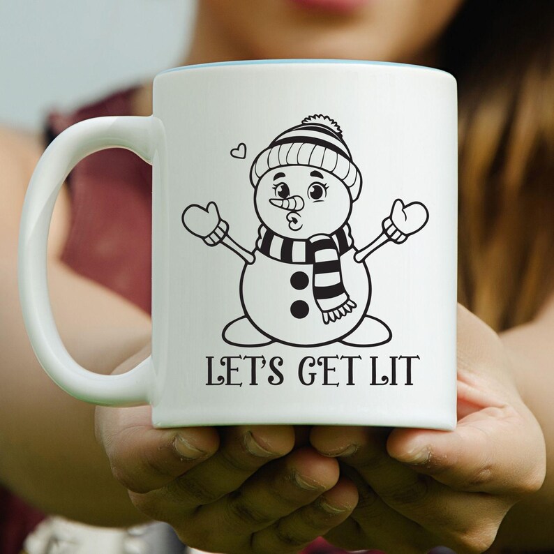 Op de afbeelding: Witte keramische mok met een cartoon sneeuwpop ontwerp, met een gestreepte sjaal, hoed en wanten. De mok heeft de tekst "LET'S GET LIT" in het zwart. De mok wordt in de handen van een persoon gehouden.