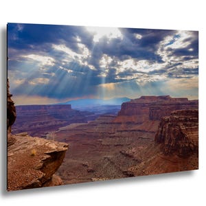 Puede incluir: Fotografía de paisaje de un cañón con formaciones rocosas rojas bajo un cielo dramático. La luz del sol atraviesa las nubes, iluminando el cañón y creando una sensación de profundidad y grandeza. La imagen captura la belleza natural del suroeste americano.