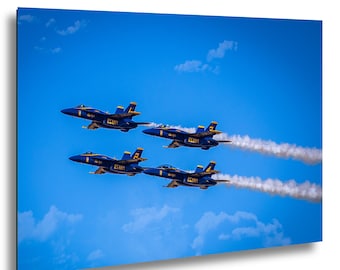 Puede incluir: Cuatro aviones de combate azul y amarillo de la Marina de los EE. UU. volando en formación contra un cielo azul brillante con nubes blancas. Los aviones dejan estelas de vapor blancas.
