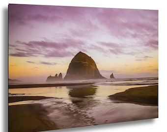 May include: A scenic landscape photograph of a beach at sunset. The image features a large rock formation in the center, with smaller rocks and the ocean in the foreground. The sky is filled with pink and purple clouds, and the water reflects the colors of the sunset.